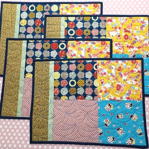Puede incluir: Un juego de cuatro manteles individuales acolchados con un diseño de patchwork. Cada mantel individual presenta una combinación de telas estampadas en azul marino, amarillo, rojo y marrón. Los manteles individuales son rectangulares y tienen un borde azul oscuro.