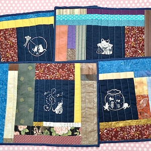 May include: Four handmade quilted placemats with a navy blue center panel featuring white cat illustrations. The placemats have a patchwork design with various fabric patterns and colors, including floral and geometric prints, and are bordered with a dark blue fabric.