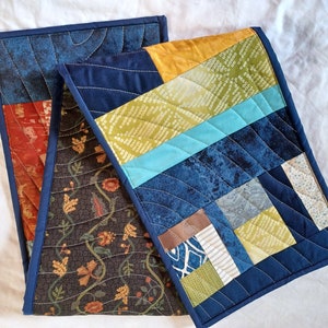 May include: Two quilted table runners, one with a dark brown floral pattern and the other with a patchwork design of blue, green, yellow, and white fabrics.