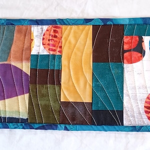 May include: A rectangular, quilted table runner with a vibrant patchwork design. The runner features vertical panels in various colors, including teal, gold, purple, and orange, with curved stitching. A blue patterned border frames the colorful center.