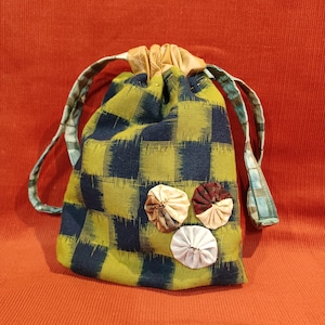 May include: A drawstring bag with a green and navy blue checkered pattern. The bag has a tan leather-like top and a green and blue patterned drawstring. Three fabric flower embellishments are sewn on the front.