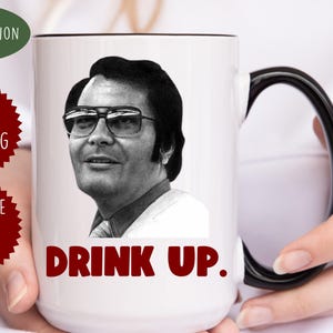 May include: White ceramic mug with a black handle. The mug features a black and white photo of a man wearing glasses and the text "Drink Up."