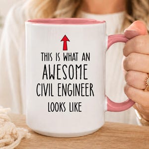 May include: White ceramic mug with a pink interior and handle. The mug features the text "THIS IS WHAT AN AWESOME CIVIL ENGINEER LOOKS LIKE" in black, with a red arrow pointing upwards. The mug is held by a person.