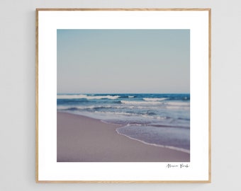 May include: A framed print of a beach scene. The image features a sandy shore, gentle waves, and a light blue sky. The artwork is in a wooden frame, creating a serene and calming aesthetic. The print is signed in the bottom right corner.