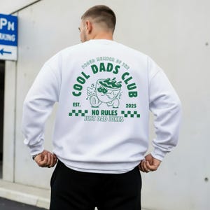 May include: White sweatshirt featuring a green graphic on the back. The graphic includes the text "COOL DADS CLUB", a cartoon coffee cup, and the phrase "NO RULES JUST DAD JOKES". The sweatshirt has a crew neck design.