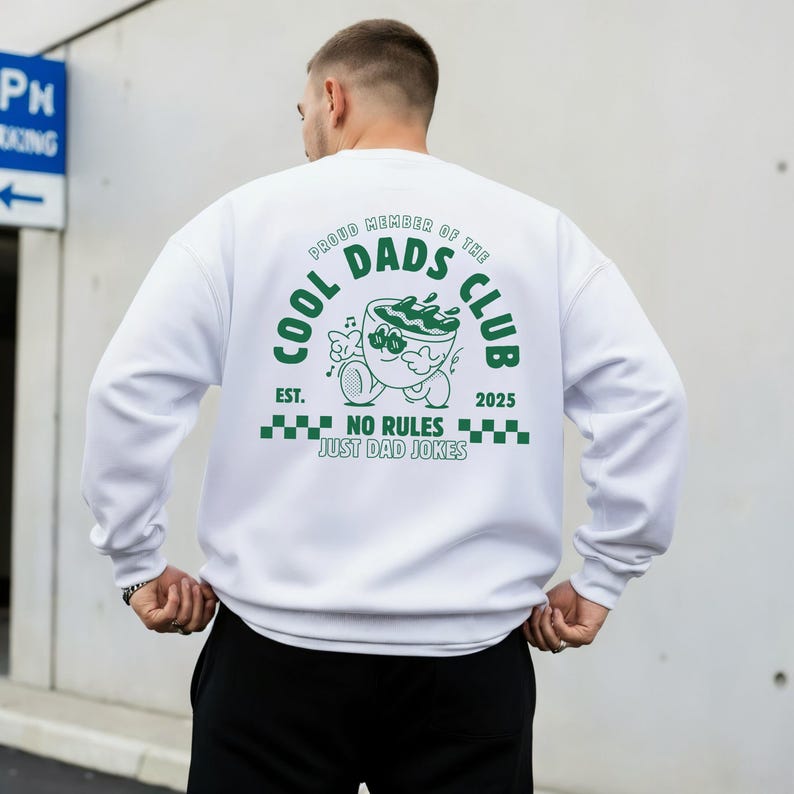 May include: White sweatshirt featuring a green graphic on the back. The graphic includes the text "COOL DADS CLUB", a cartoon coffee cup, and the phrase "NO RULES JUST DAD JOKES". The sweatshirt has a crew neck design.