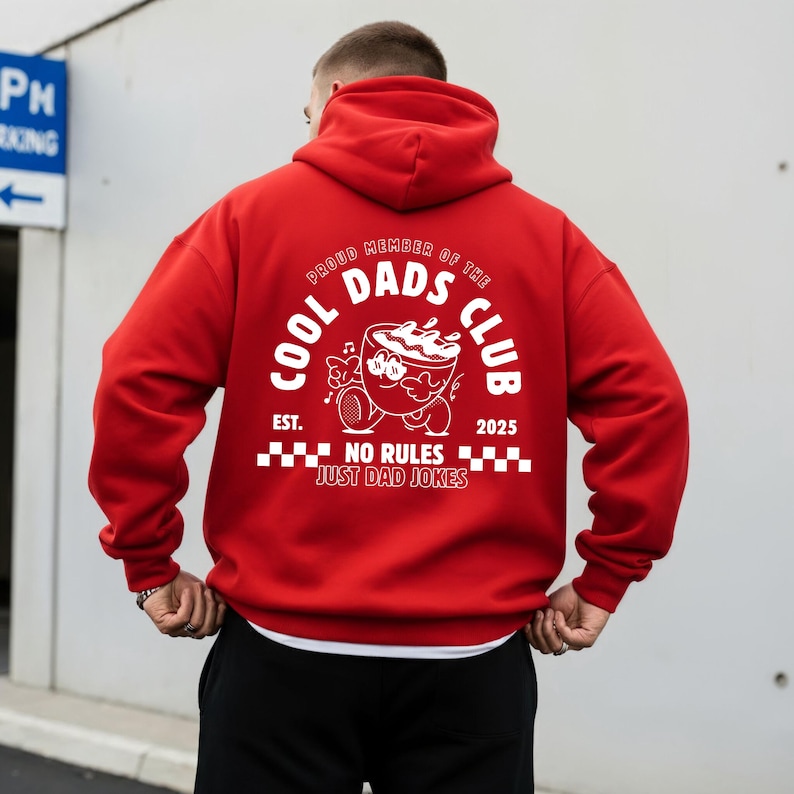May include: A red hoodie featuring a white graphic on the back. The graphic says "COOL DADS CLUB" with a cartoon coffee cup character and the text "NO RULES JUST DAD JOKES". The hoodie has a drawstring hood.