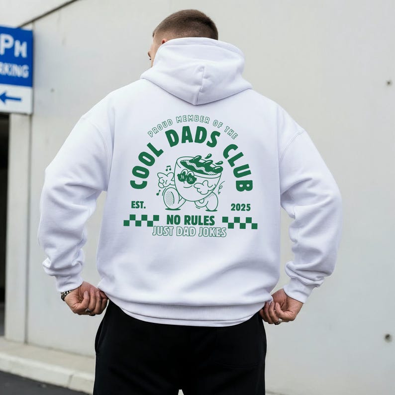 May include: White hoodie featuring a green graphic on the back. The graphic says "COOL DADS CLUB" with a cartoon design and the text "NO RULES JUST DAD JOKES". The hoodie has a hood and is made of a soft material.