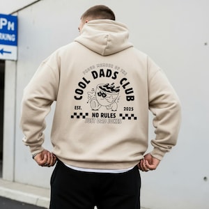 May include: Beige hoodie with a hood and long sleeves. The back features a black graphic that reads "COOL DADS CLUB" with a cartoon coffee cup design and the text "NO RULES JUST DAD JOKES". The hoodie is a neutral colour.