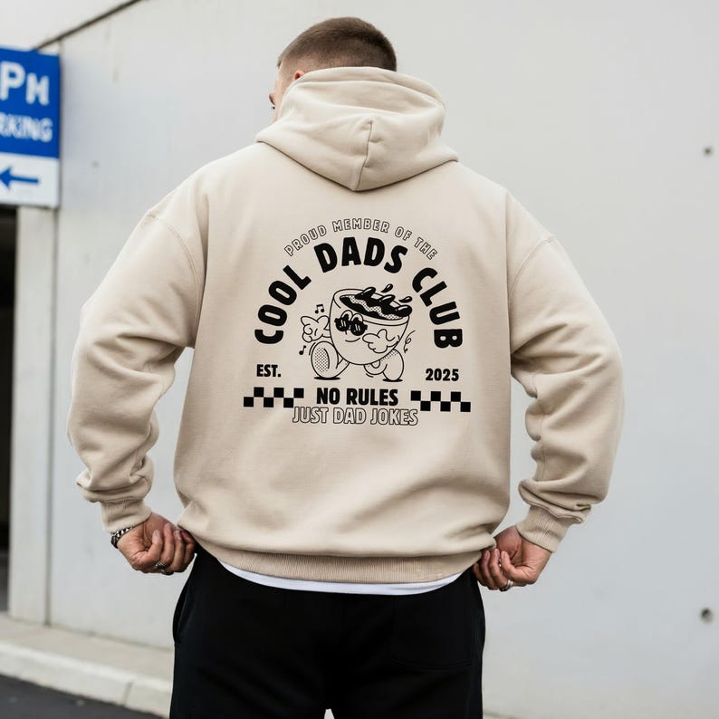 May include: Beige hoodie with a hood and long sleeves. The back features a black graphic that reads "COOL DADS CLUB" with a cartoon coffee cup design and the text "NO RULES JUST DAD JOKES". The hoodie is a neutral colour.
