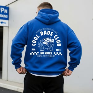 May include: A vibrant blue hoodie featuring a white graphic on the back. The design includes the text "COOL DADS CLUB" and a cartoon bowl character. The phrase "NO RULES JUST DAD JOKES" is also printed on the hoodie.