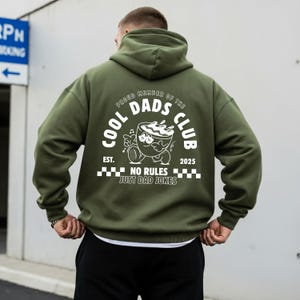 May include: An olive green hoodie featuring a white graphic on the back. The graphic includes the text "COOL DADS CLUB" and a cartoon coffee cup character. The phrase "NO RULES JUST DAD JOKES" is also printed.