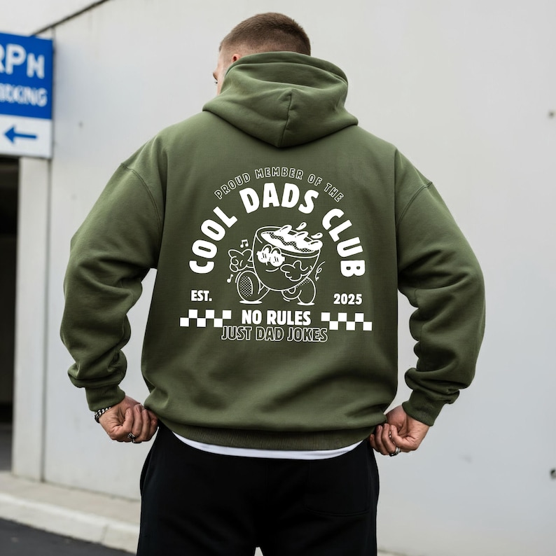 May include: An olive green hoodie featuring a white graphic on the back. The graphic includes the text "COOL DADS CLUB" and a cartoon coffee cup character. The phrase "NO RULES JUST DAD JOKES" is also printed.