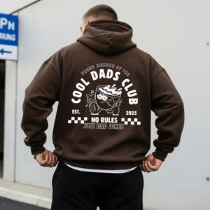 May include: A brown hoodie featuring a white graphic on the back. The graphic includes the text "COOL DADS CLUB" and a cartoon coffee cup character. The phrase "NO RULES JUST DAD JOKES" is also printed.