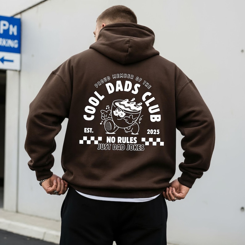May include: A brown hoodie featuring a white graphic on the back. The graphic includes the text "COOL DADS CLUB" and a cartoon coffee cup character. The phrase "NO RULES JUST DAD JOKES" is also printed.