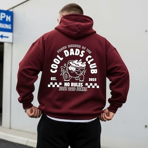 May include: A maroon hoodie featuring a white graphic on the back. The graphic includes the text "COOL DADS CLUB" and a cartoon coffee cup character. The phrase "NO RULES JUST DAD JOKES" is also printed.