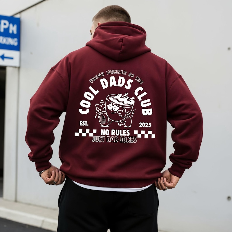 May include: A maroon hoodie featuring a white graphic on the back. The graphic includes the text "COOL DADS CLUB" and a cartoon coffee cup character. The phrase "NO RULES JUST DAD JOKES" is also printed.