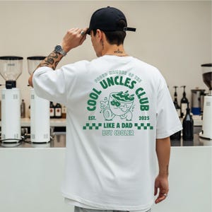May include: White t-shirt with a green graphic on the back that reads "COOL UNCLES CLUB" with a cartoon coffee cup design. The text "LIKE A DAD BUT COOLER" is also printed on the shirt. The shirt is being worn by a person.