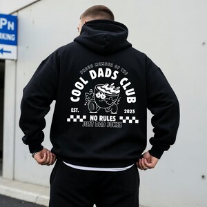 May include: Black hoodie featuring a white graphic on the back. The graphic includes the text "COOL DADS CLUB" and a cartoon coffee cup character. The words "PROUD MEMBER OF THE DADS CLUB" are arched above the graphic, with "EST. 2025" below.