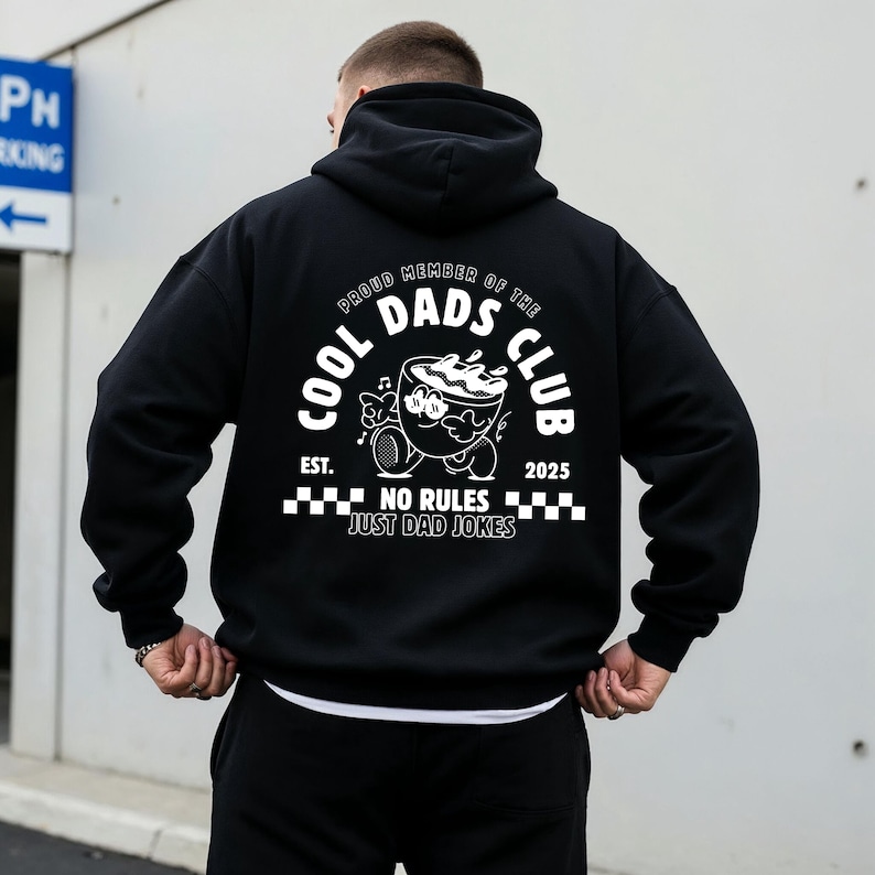 May include: Black hoodie featuring a white graphic on the back. The graphic includes the text "COOL DADS CLUB" and a cartoon coffee cup character. The words "PROUD MEMBER OF THE DADS CLUB" are arched above the graphic, with "EST. 2025" below.