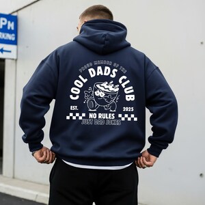 May include: A navy blue hoodie featuring a white graphic on the back. The graphic includes the text "COOL DADS CLUB" and a cartoon coffee cup design. The text "NO RULES JUST DAD JOKES" is also present. The hoodie has a hood and a relaxed fit.