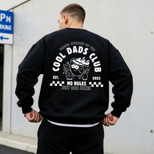 May include: Black sweatshirt featuring a white graphic on the back. The graphic includes the text "COOL DADS CLUB" and a cartoon coffee cup. The phrase "NO RULES JUST DAD JOKES" is also present. The sweatshirt has a crew neck.