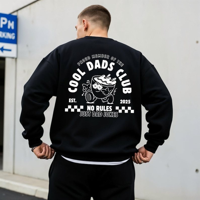 May include: Black sweatshirt featuring a white graphic on the back. The graphic includes the text "COOL DADS CLUB" and a cartoon coffee cup. The phrase "NO RULES JUST DAD JOKES" is also present. The sweatshirt has a crew neck.