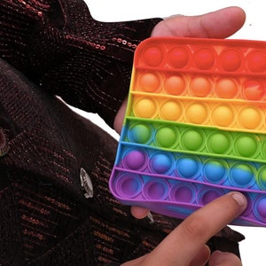 May include: A rainbow-colored pop-it fidget toy with a square shape and multiple rows of bubbles. The toy is made of silicone and is designed to be squeezed and popped.