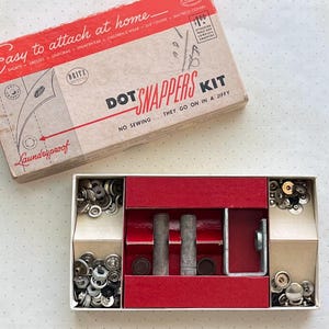 May include: A vintage Dot Snappers kit in a rectangular cardboard box with red and white graphics. The box lid features the text Easy to attach at home and Dot Snappers Kit. Inside, the tray contains metal snap fasteners and tools.
