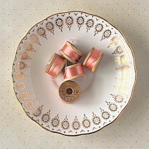 May include: A white porcelain saucer with a gold and white floral pattern, containing four spools of pink thread and a wooden spool of thread with the text "TWIST" and "25c".
