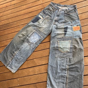 Peut inclure: Jean en denim bleu clair avec un design patchwork. Le jean présente de multiples patchs en denim de différentes nuances de bleu, avec une étiquette rouge Levi's sur la poche arrière. Le jean est posé sur une surface en bois.