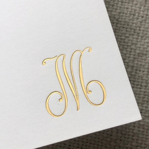 May include: A white card with a gold foil monogram of the letter "M" in a cursive script.
