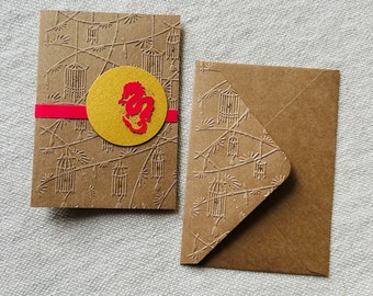 May include: A brown card with a red stripe and a gold circle with a red dragon inside. The card has a pattern of cages and branches. The card is next to a brown envelope with the same pattern.