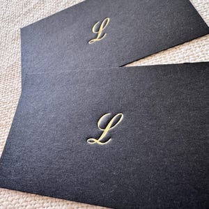 May include: Two black cards with a gold letter L on each. The cards are on a white textured surface.