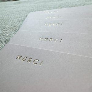 May include: Close-up of several light gray cards with the word "MERCI" embossed in gold. The cards are arranged in a slightly fanned-out pattern, with the text clearly visible. The background is a soft, textured fabric.