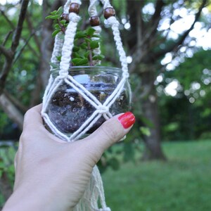 May include: A white macrame plant hanger with wooden beads holds a glass jar with soil and a small green plant. The hanger is being held by a hand with red nail polish.