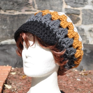 K&ouml;nnte beinhalten: Eine gestrickte M&uuml;tze mit schwarzen, grauen und wei&szlig;en Streifen und einem Band aus braunen geh&auml;kelten Quadraten am Rand.