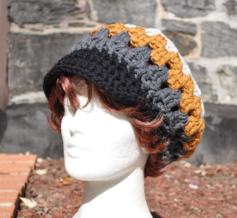 K&ouml;nnte beinhalten: Eine gestrickte M&uuml;tze mit schwarzen, grauen und wei&szlig;en Streifen und einem Band aus braunen geh&auml;kelten Quadraten am Rand.