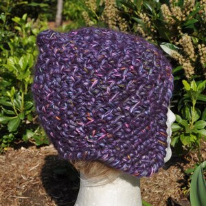 May include: Purple crocheted hat with a slouchy, beanie style and a long, pointed ear flap on one side. The hat is made with a variegated yarn that includes shades of purple, pink, and blue.