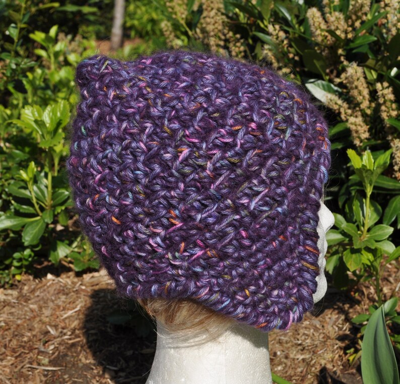 May include: Purple crocheted hat with a slouchy, beanie style and a long, pointed ear flap on one side. The hat is made with a variegated yarn that includes shades of purple, pink, and blue.