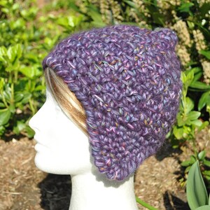 May include: Purple crocheted hat with a slouchy fit and ear flaps. The hat is made with a variegated yarn that has shades of purple, pink, and blue.