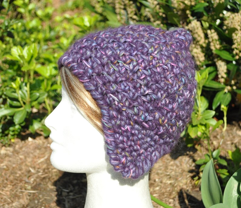 May include: Purple crocheted hat with a slouchy fit and ear flaps. The hat is made with a variegated yarn that has shades of purple, pink, and blue.