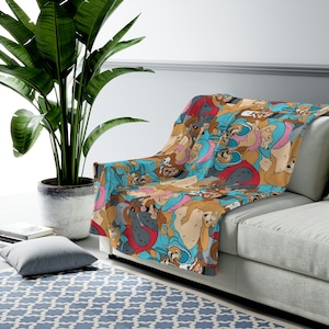 May include: A colorful throw blanket featuring a repeating pattern of cartoon dogs in various poses and outfits. The blanket is draped over a gray sofa, with a large green plant in the background. The blanket's design includes shades of brown, blue, pink, and red.