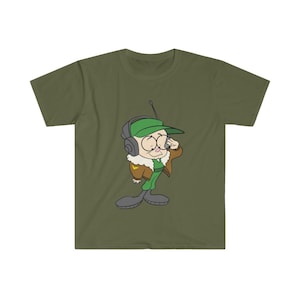 以下が含まれることがあります： オリーブグリーンのTシャツ。緑の帽子、ヘッドフォン、茶色のジャケットを着た漫画のキャラクターが描かれています。キャラクターは大きな目を持ち、耳に触れています。デザインはシャツの中央に配置されています。