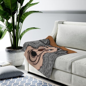 Könnte beinhalten: Eine grau-braune Decke mit einem Cartoon-Wolf-Design, drapiert über einem weißen Sofa. Die Decke hat eine Kunstfellstruktur und einen braunen Rand. Eine große Topfpflanze und ein blau gemusterter Teppich befinden sich im Hintergrund.
