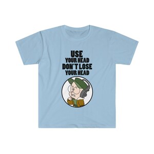 以下が含まれることがあります： 「USE YOUR HEAD DON'T LOSE YOUR HEAD」の黒い文字が入った水色のTシャツ。緑の帽子、メガネ、ヘッドホンをつけた漫画のキャラクターが白い円の中に入っています。キャラクターは茶色のジャケットを着ています。