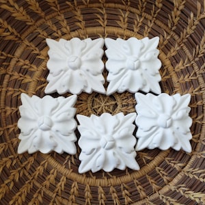 May include: Five white plaster floral embellishments with a raised center and a square shape. The embellishments are arranged in a circle on a woven basket surface.