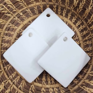 May include: Three white ceramic square tiles with a hole in the top corner. The tiles are on a woven brown basket.