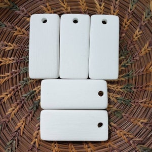 May include: A collection of six rectangular, white painted wooden tags with a small hole drilled at the top. The tags are arranged on a woven, brown and green textured surface.