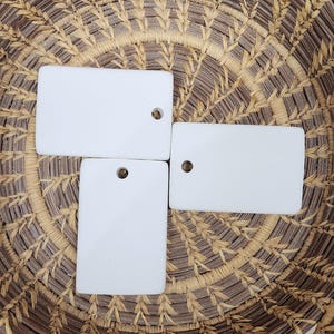 May include: Three rectangular white ceramic tags with smooth surfaces and rounded corners. Each tag features a single circular hole punched near one edge, designed for hanging or attachment. The tags are plain, solid white, and matte in finish.